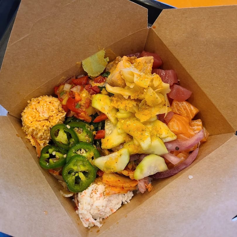 Colorful poke bowl with salmon, tuna, jalapenos, cucumbers, and rice in a cardboard box.