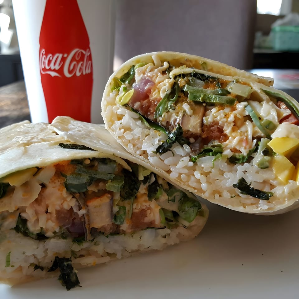 Two halves of a poke sushi burrito filled with rice, fish, and vegetables, with a Coca-Cola cup.