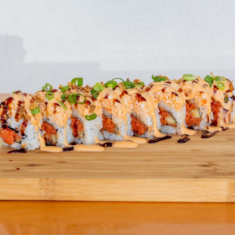 A row of sushi rolls with orange sauce, dark drizzle, and green onions on a wooden board.